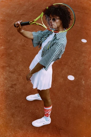 Woman in white pleated tennis skirt holding a racket over her head on a clay tennis court