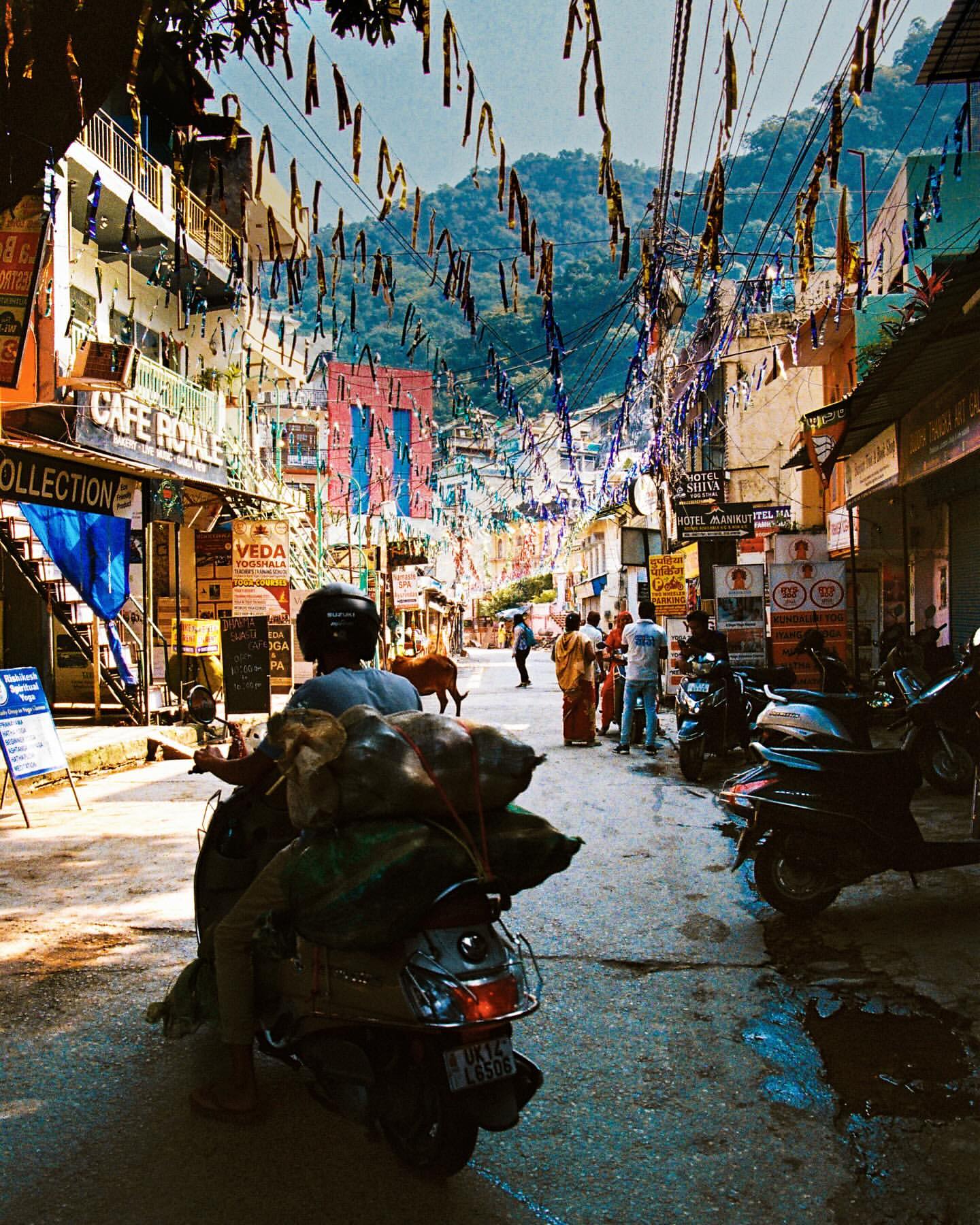 Street view in India showcases a motorcyclist on a scooter, pedestrians, and colorful buildings under a sky strung with decorations.