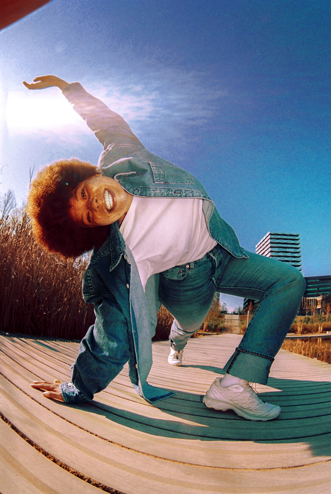 Smiling person in double denim holding a yoga pose on a wooden boardwalk under a clear blue sky