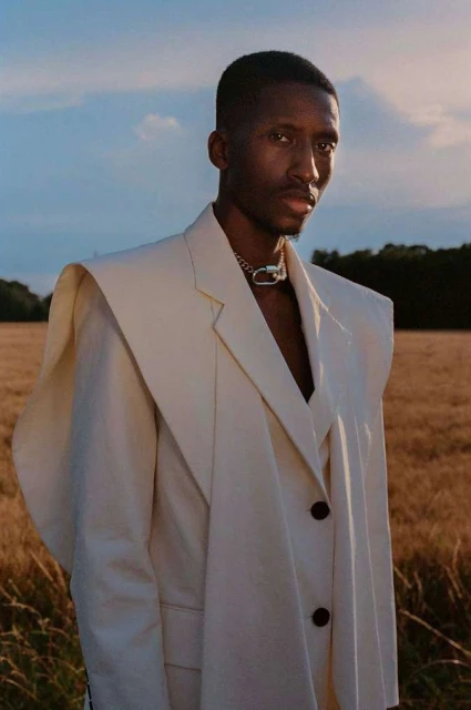 Portrait of a Black male model in a cream-colored blazer with a statement silver chain against a blurred wheat field background.