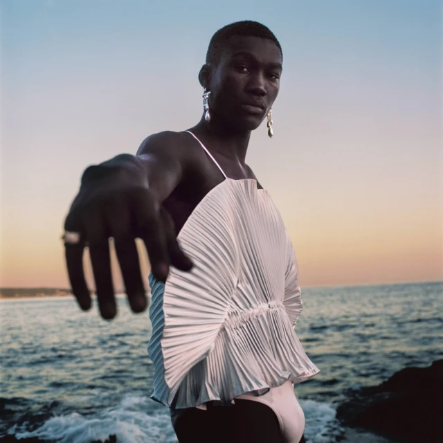 A person with dark skin, wearing a white pleated top and earrings, stands by the ocean in the early morning, with arm extended.