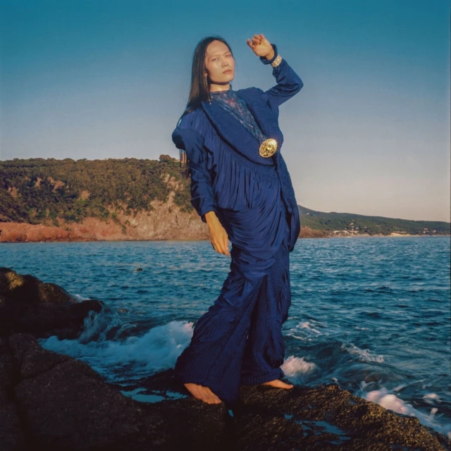 Woman in flowing blue dress poses barefoot on rocks by the ocean under a clear sky, with coastal vegetation visible in the background.