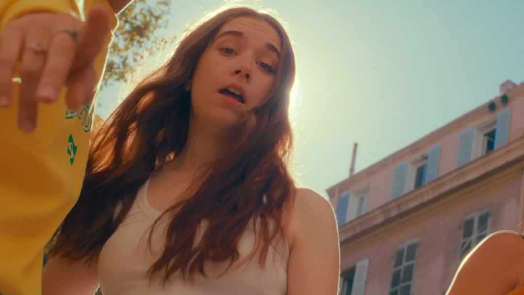 Close-up of a young adult female in a white tank top, glancing up at the camera under bright sunlight with a building backdrop.