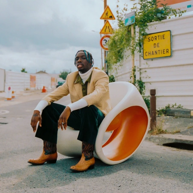 A young black man sits in a white egg chair on a street with road signs, wearing a tan blazer, white turtleneck, dark pants and cowboy boots.