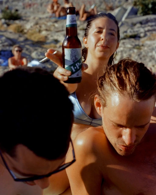 Woman holds La Chappelle IPA bottle on sunny beach with people relaxing; close-up shot, eye-level view, sunlit environment, casual attire.
