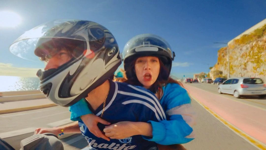 Motorcycle ride with a couple wearing helmets, hugging, and driving down a coastal road on a sunny day with parked cars and ocean view.