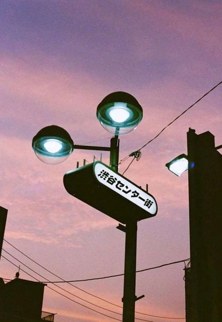Evening view of a Japanese street sign in Shibuya Center district with streetlights against a colorful pink and purple sky.