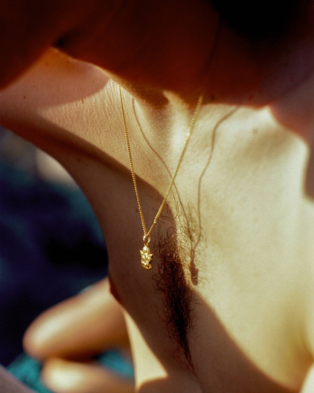 Close-up of an adult wearing a gold necklace with a pendant. The person is lit by natural light, with shadows adding contrast to the skin.