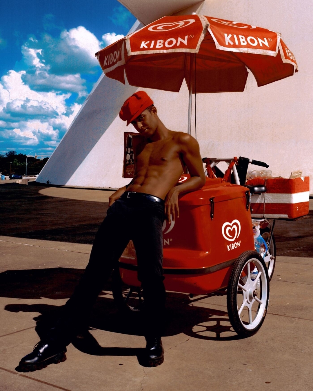 A shirtless adult male leans against a red Kibon ice cream cart under an umbrella, wearing black pants and boots, with a modern building behind.