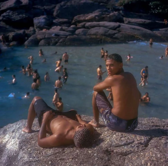 Two shirtless men on a rock near a beach filled with many people in the water in an outdoor setting with natural lighting.