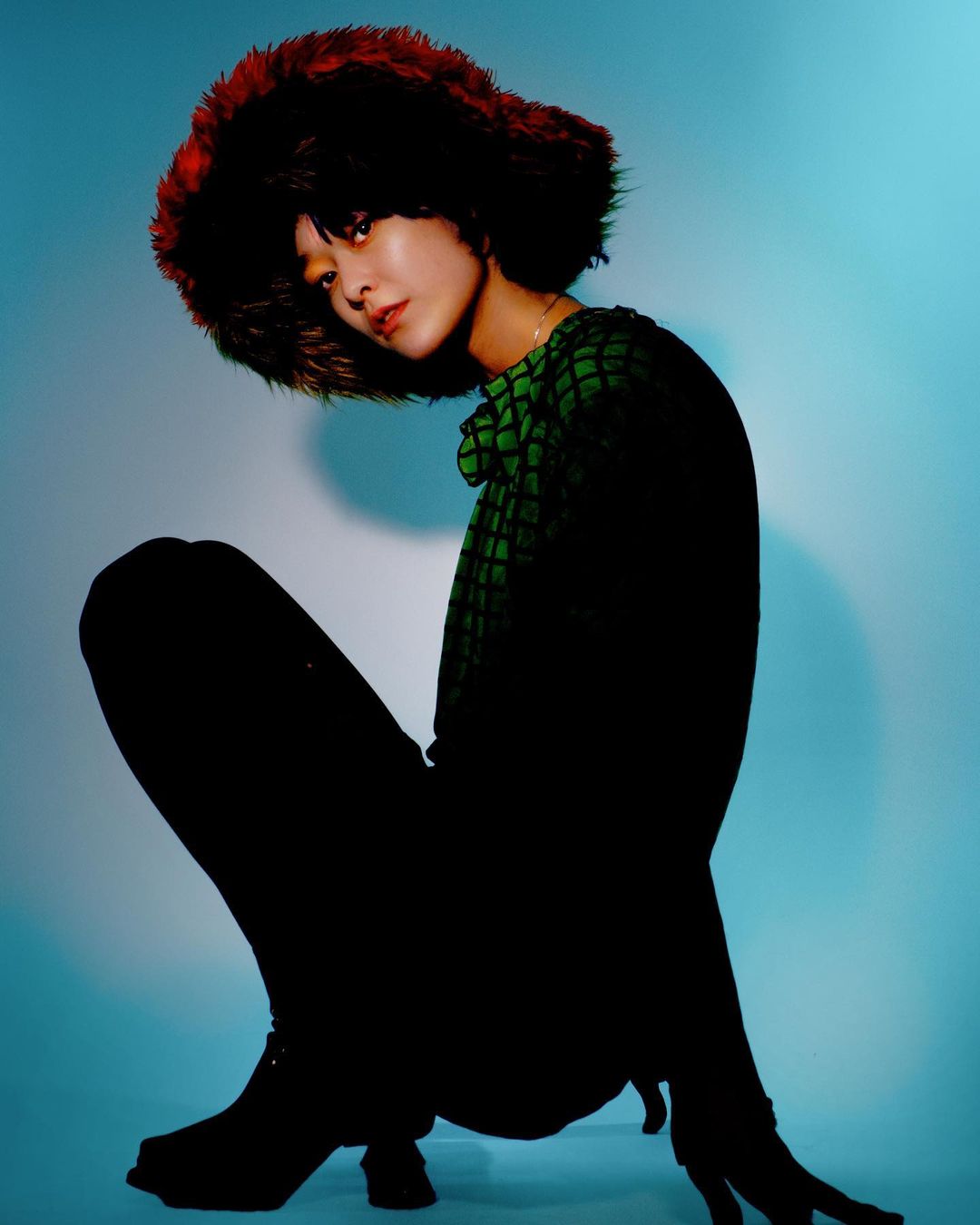 Woman with an auburn fur hat poses in a dark top and pants, crouching with a blue gradient background in a fashion shot.