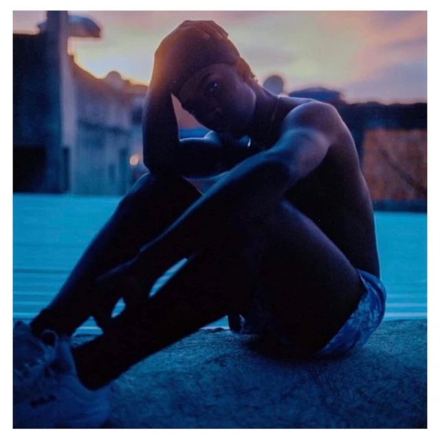 Man in hat sitting on concrete, wearing shorts and sneakers. Silhouette against sunset sky with architectural details visible.