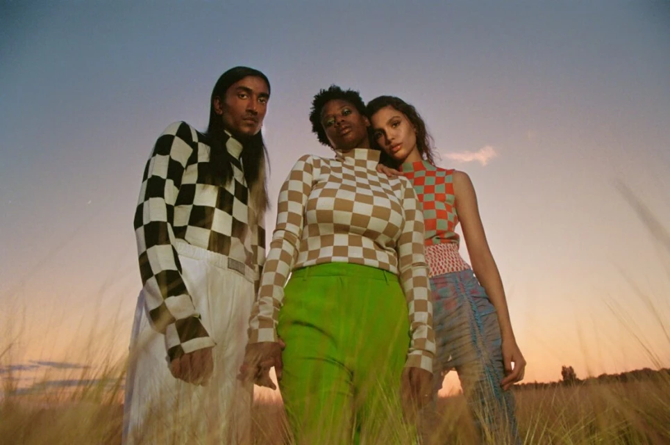 Three fashion models pose in a field at dusk, wearing checkered knitwear tops and stylish trousers, with a warm, muted color palette.