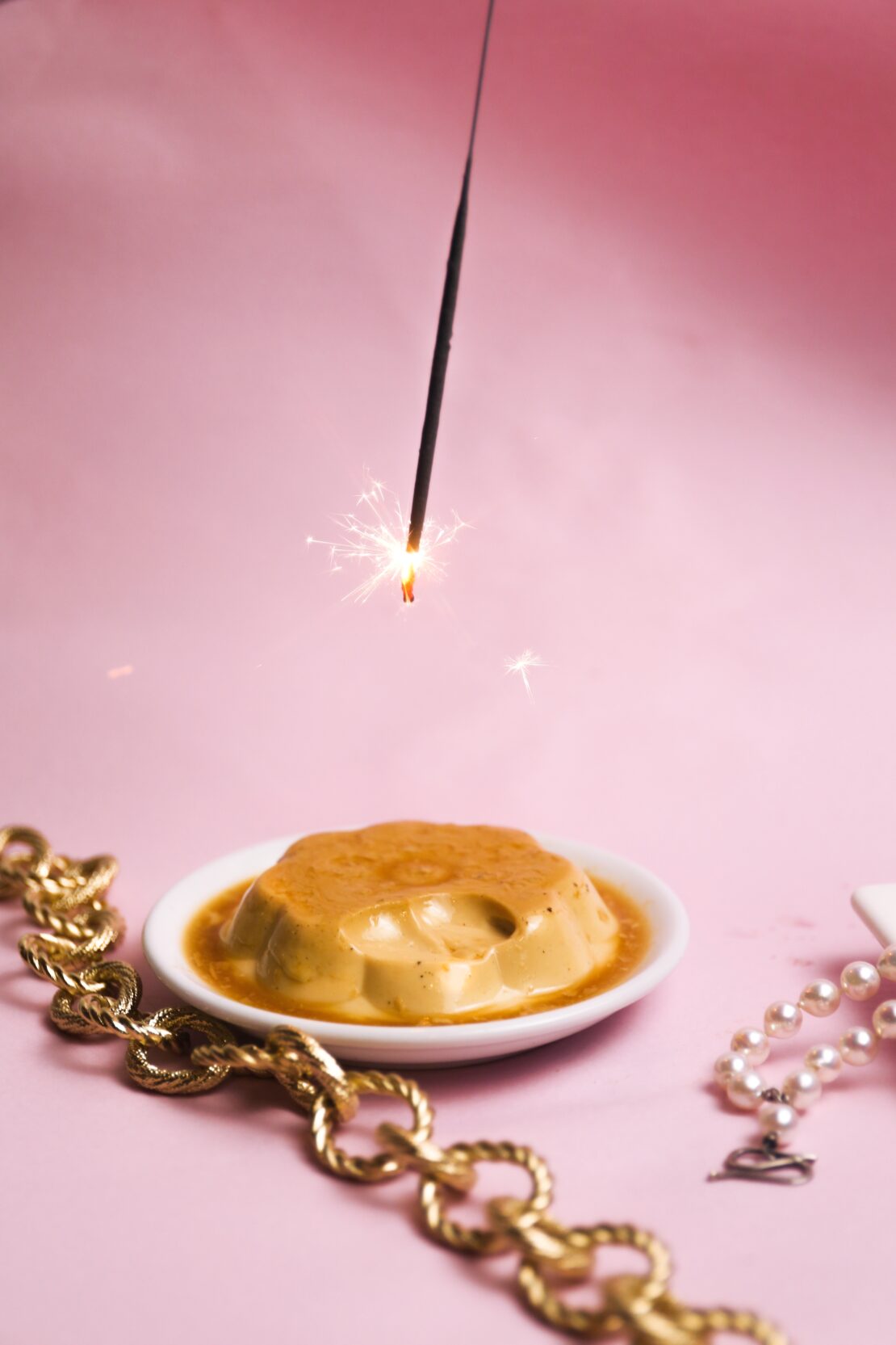 Still life food shot of a caramel flan dessert on a white plate, gold jewelry, sparkler, against a light pink background.