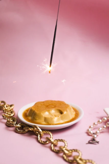Still life food shot of a caramel flan dessert on a white plate, gold jewelry, sparkler, against a light pink background.