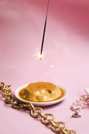 Still life food shot of a caramel flan dessert on a white plate, gold jewelry, sparkler, against a light pink background.