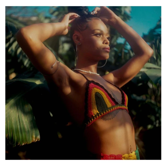 Young woman poses outdoors in a crochet bikini top with green leafy background. She is arranging her hair in an outdoor setting.