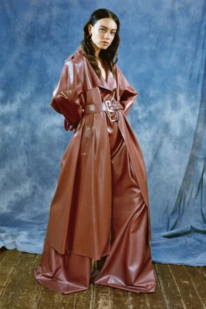 Model in brown belted trench coat and matching trousers standing against a textured blue backdrop.