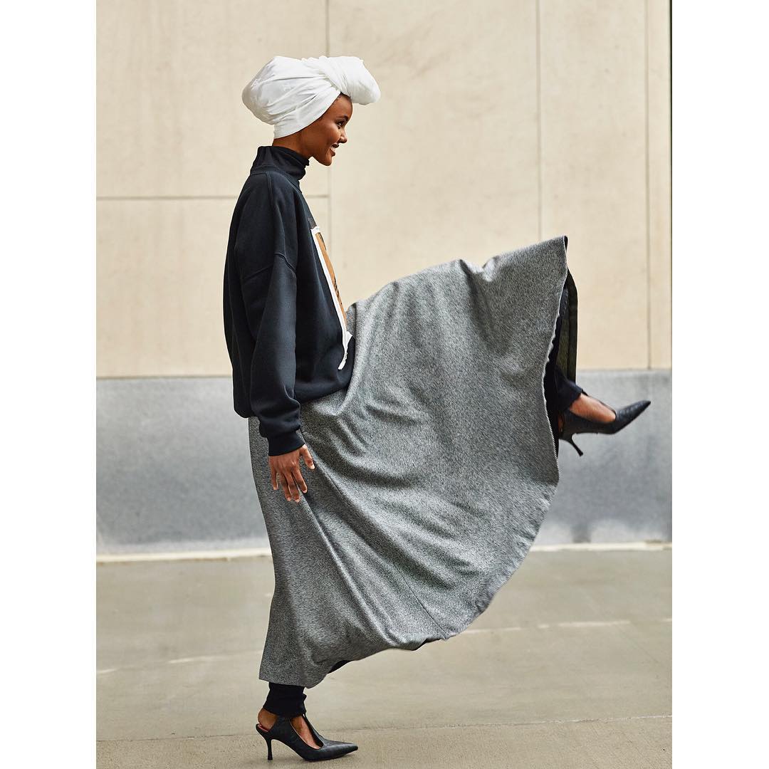 A woman in a black top and grey skirt poses outdoors, lifting her leg to display a black pointed-toe heel, wearing a white head wrap.