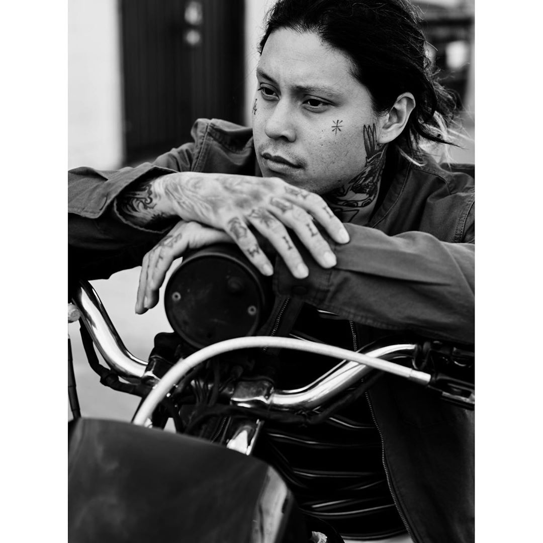 Portrait of a tattooed man leaning on a motorcycle in black and white, gazing to the side with a serious expression.
