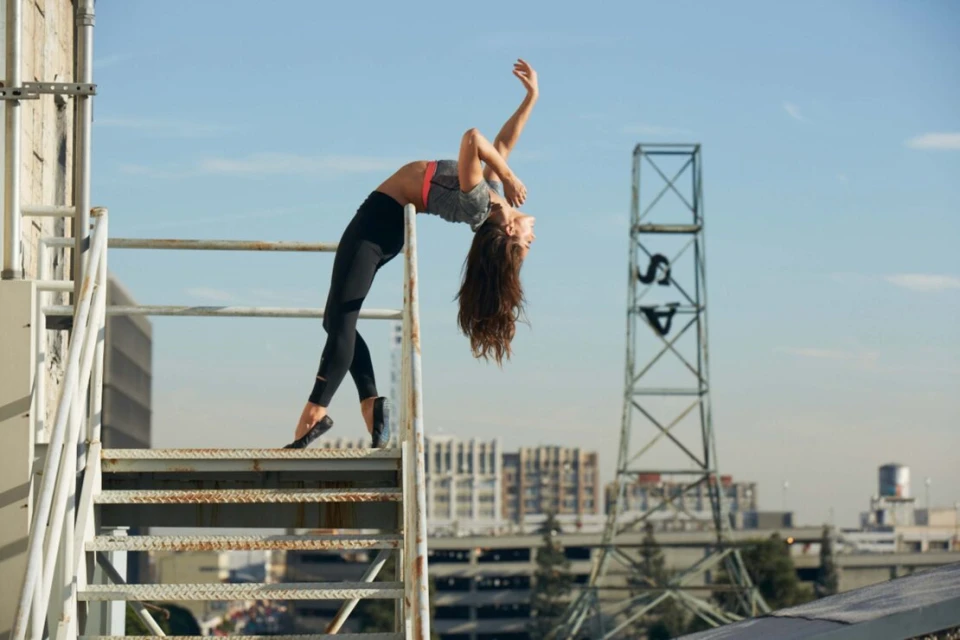 Female dancer in activewear performs a backbend on a metal staircase in an urban setting with buildings and a communications tower.