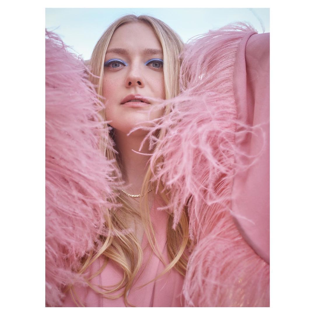 Blonde woman in pink feather-trimmed jacket with light blue eye makeup, pearl necklace, and neutral expression against a light sky.