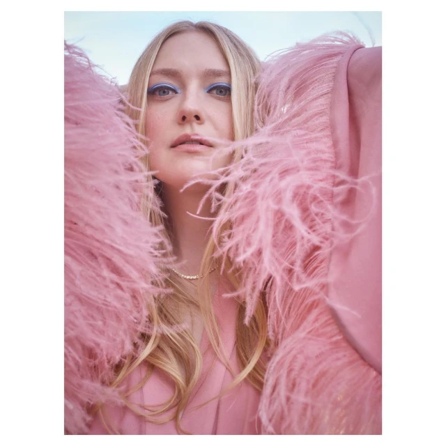 Blonde woman in pink feather-trimmed jacket with light blue eye makeup, pearl necklace, and neutral expression against a light sky.