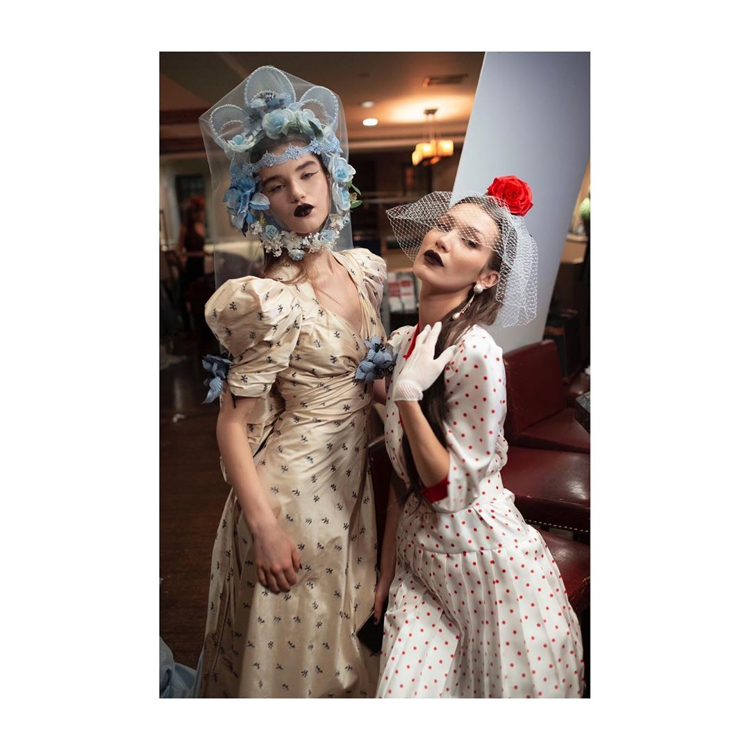 Two fashion models pose indoors wearing vintage-style dresses and floral headpieces, one in red polka dots, capturing a retro aesthetic.