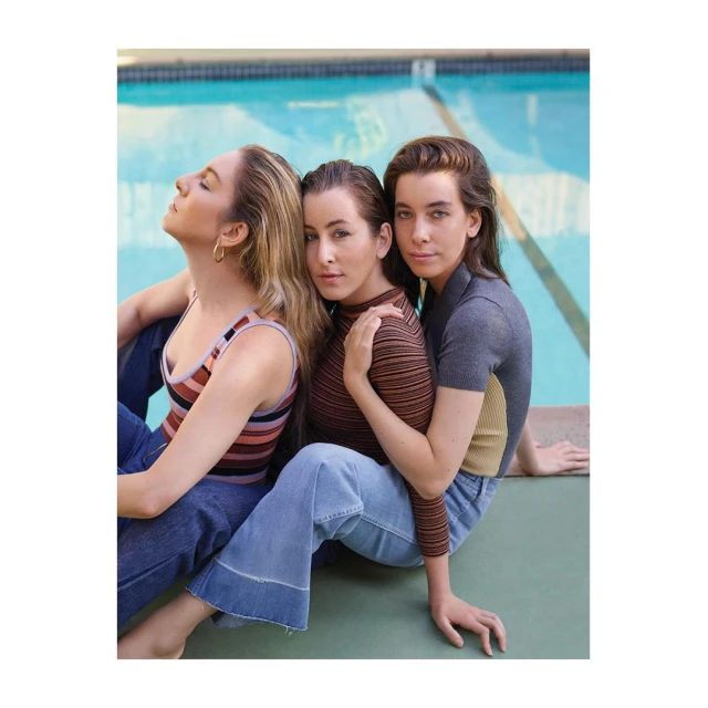 Three women sit in front of a swimming pool, wearing casual striped tops and jeans in a medium shot. Natural lighting.