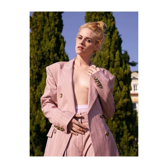 Stylish woman in a pink pinstripe suit poses outdoors against a backdrop of green trees and blue sky, exuding confidence and fashion.