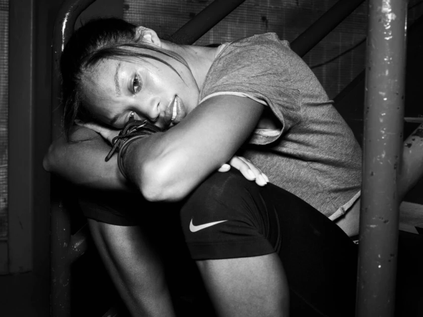 Monochrome portrait of athletic adult woman in sportswear resting head on arms with hair pulled back, sitting indoors against metal bars.
