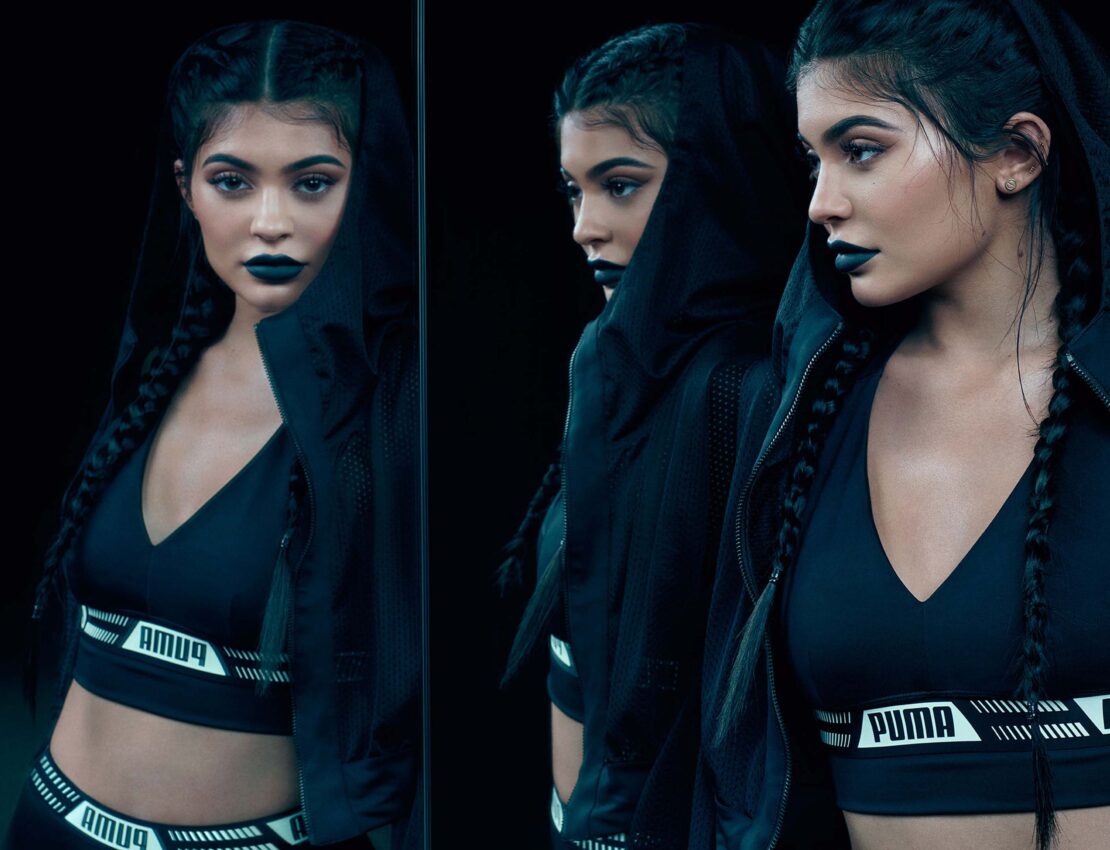 Close-up of an adult female fashion model with braided hair wearing dark sportswear with a logo, posing in front of a dark background.