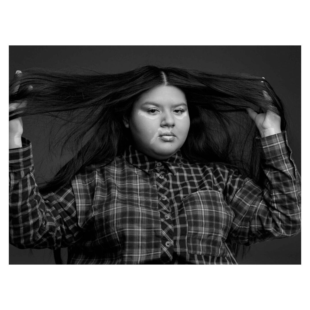 Portrait of a woman with long dark hair wearing a plaid shirt. The black-and-white photo shows her holding her hair above her head.