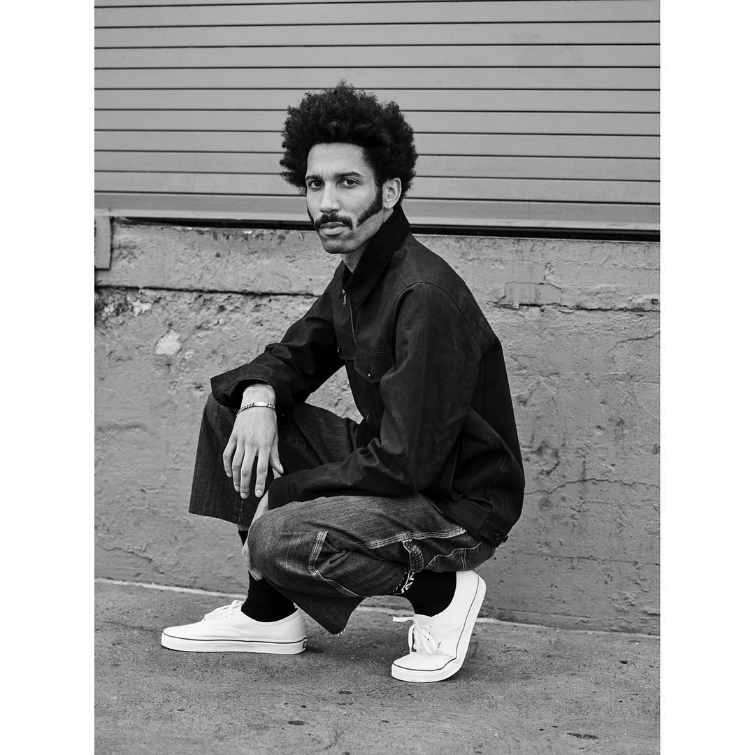 Monochrome portrait of a stylish adult male crouching outdoors in denim jacket and pants, white sneakers and dark socks against a concrete wall.