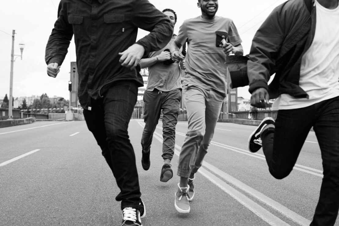 Monochrome action shot of four men running together along a wide paved road outdoors.