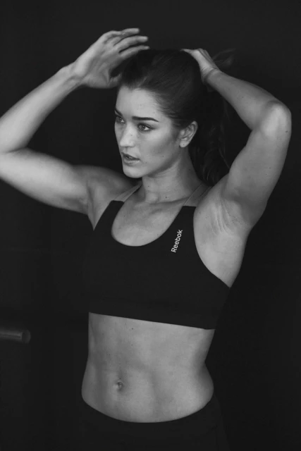 Black and white studio shot of an adult female athlete tying up her hair in sportswear against a dark background.