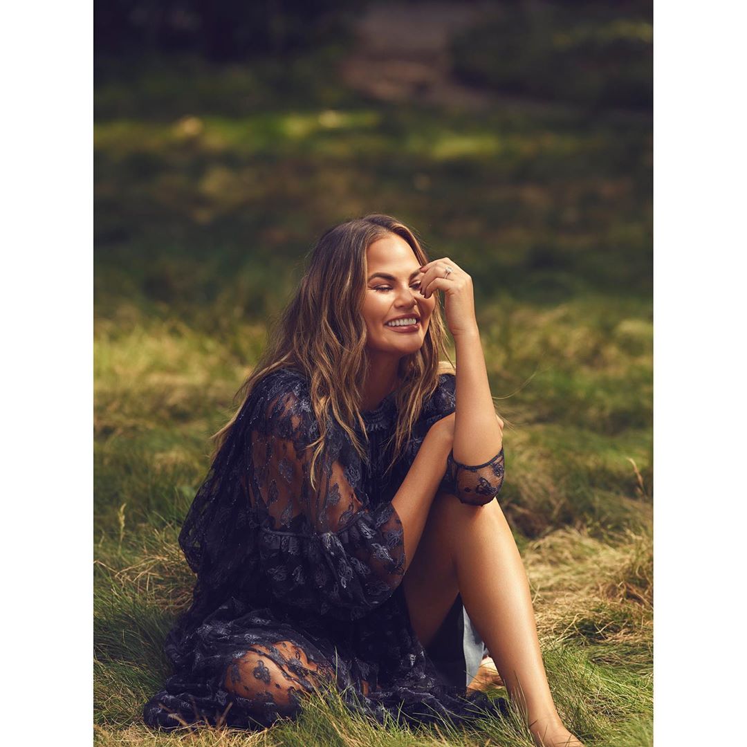 Chrissy Teigen sitting outdoors on a grassy meadow wearing a sheer dark patterned dress, smiling with hand near face.