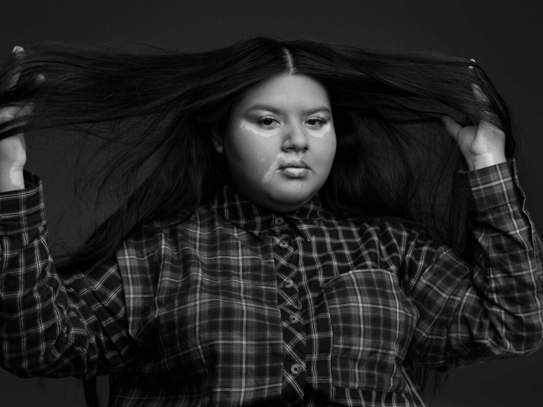 Monochrome portrait of a woman pulling her long dark hair out to the sides while wearing a plaid button-down shirt.