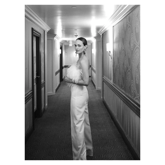 A woman in a white strapless jumpsuit with feathered details, stands in a hotel hallway with patterned walls in a black and white photograph.