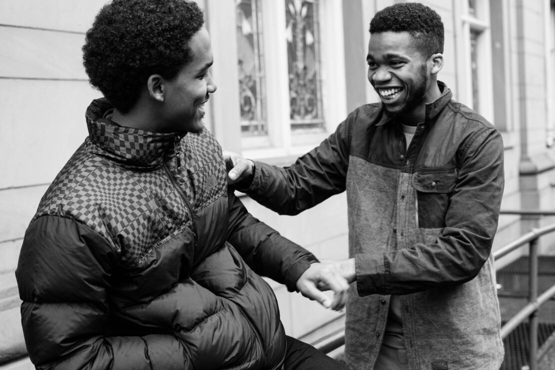 Two young African American men are outside a building, one in a puffy jacket, smiling and laughing in a black and white photograph.