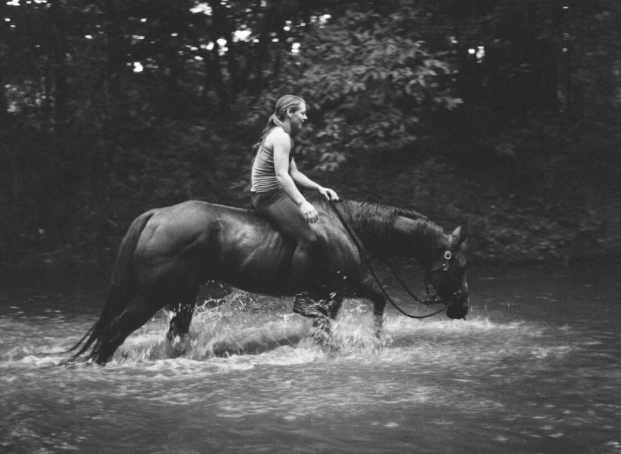 A woman rides a brown horse through shallow water with trees in the background in a black and white outdoor photograph.