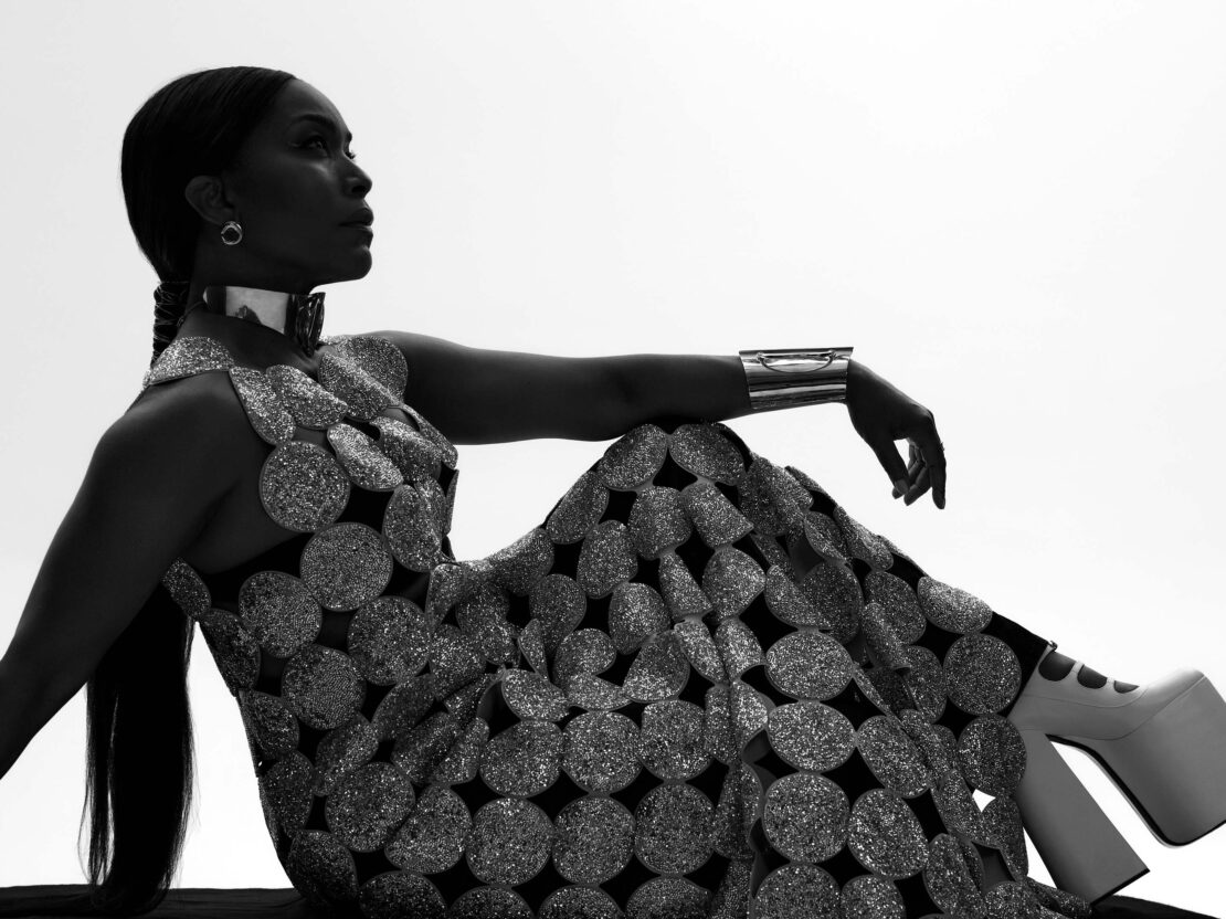 Silhouette portrait of Angela Bassett wearing a metallic disc-embellished gown, choker, cuff, and platform boots against bright sky.