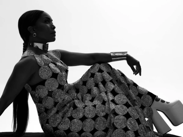 Silhouette portrait of Angela Bassett wearing a metallic disc-embellished gown, choker, cuff, and platform boots against bright sky.