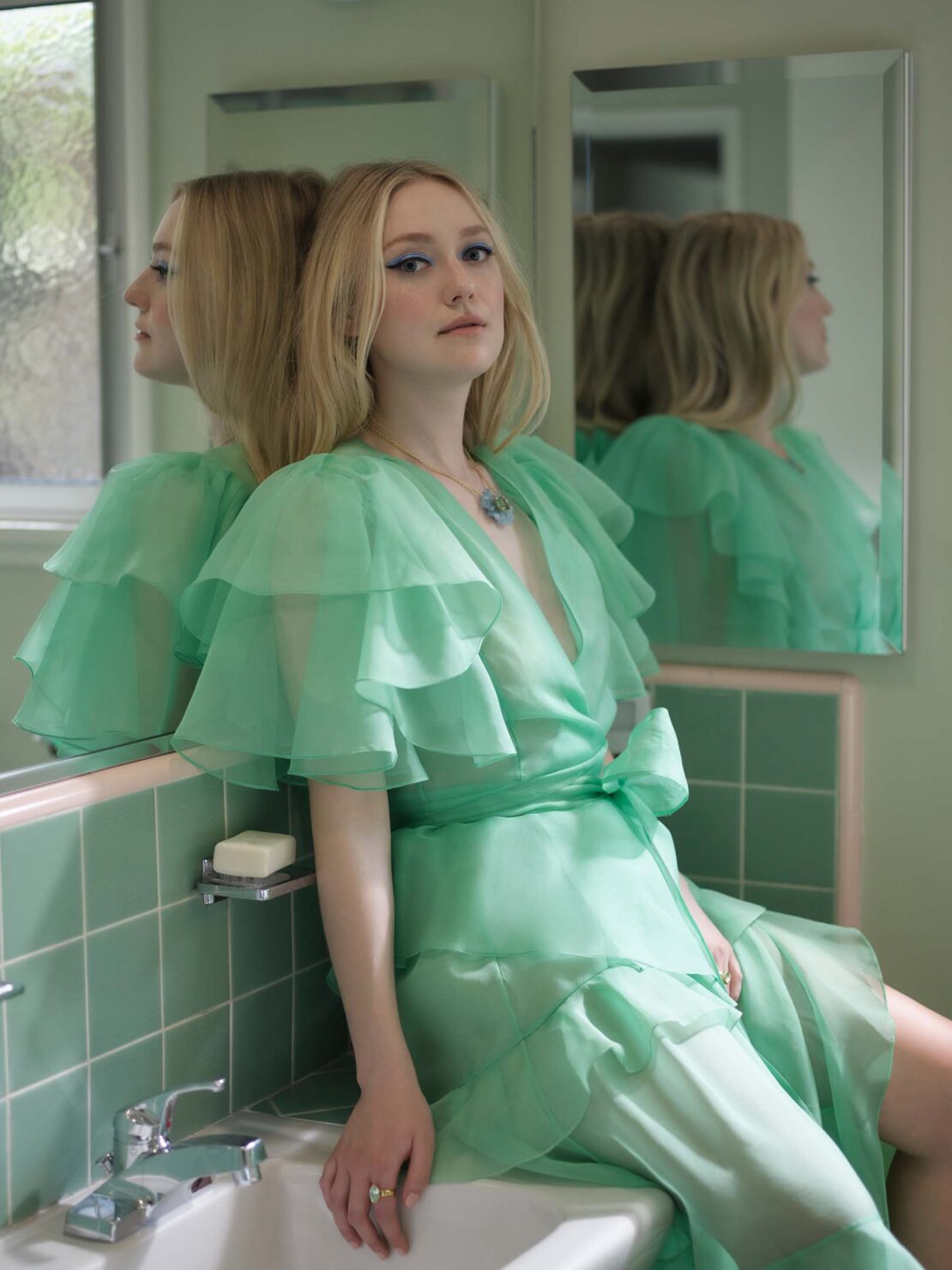 Portrait of fair-skinned woman with blonde hair wearing mint-green ruffled dress sitting on sink with reflective surface and green-tiled wall in background.
