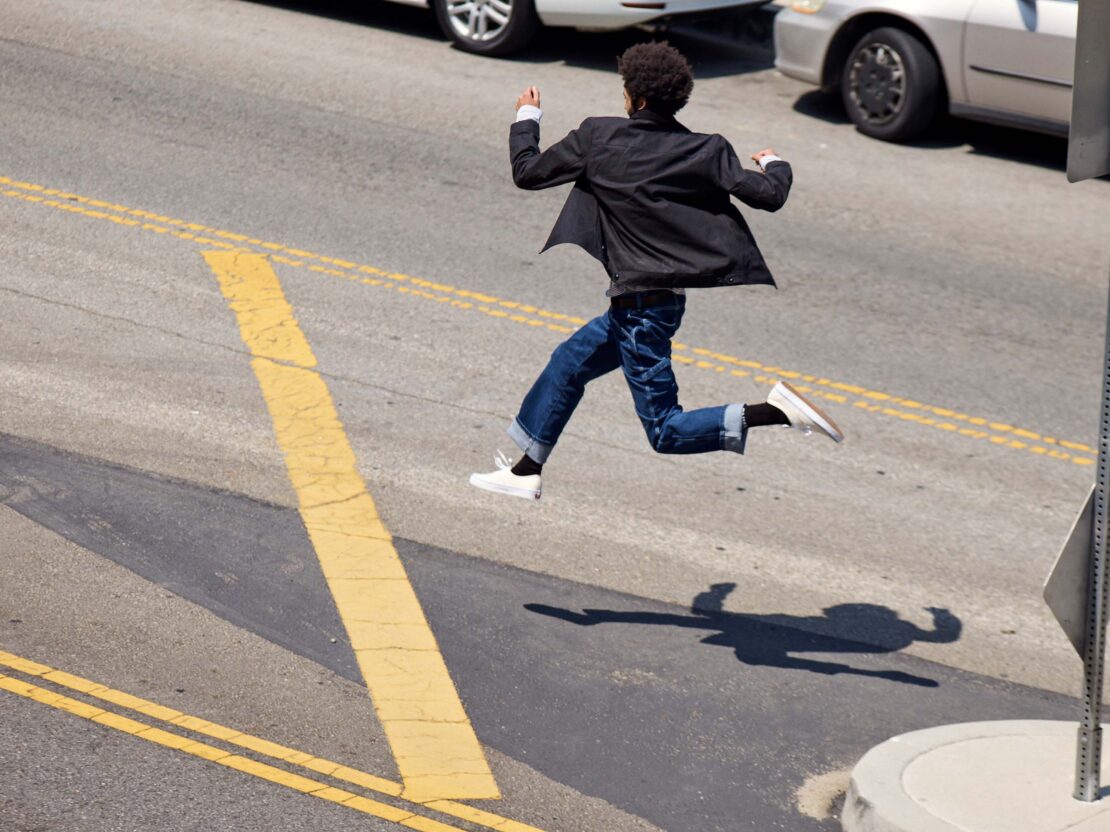 An adult male is frozen mid-air with a blazer, jeans, and sneakers; crossing a city street with yellow lines on a sunny day.