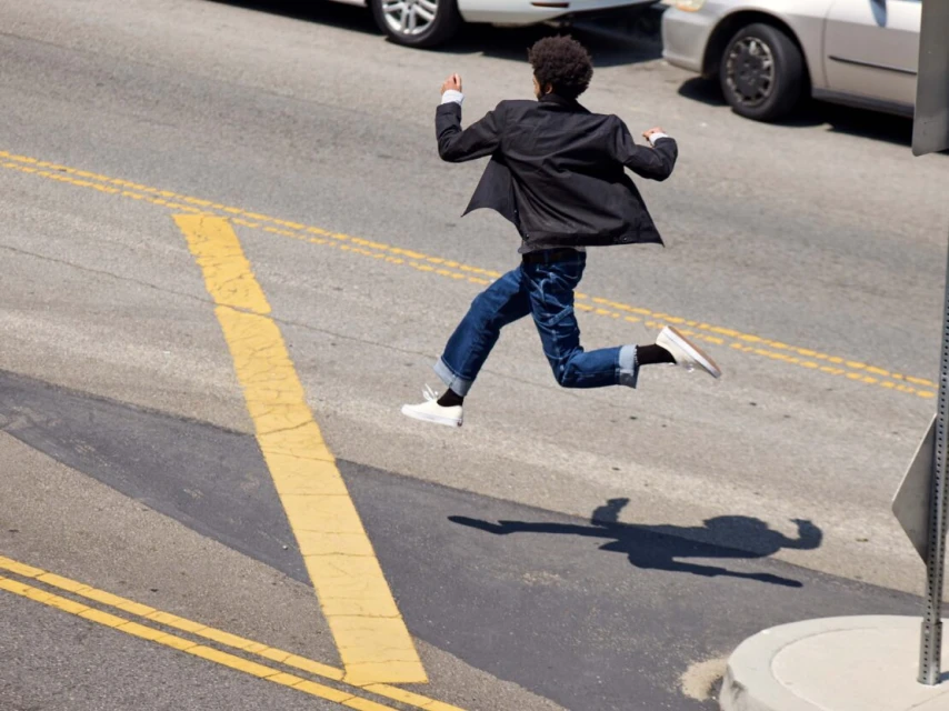 An adult male is frozen mid-air with a blazer, jeans, and sneakers; crossing a city street with yellow lines on a sunny day.