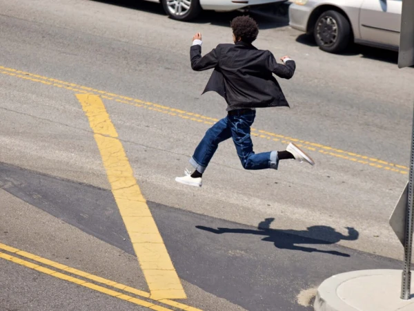 An adult male is frozen mid-air with a blazer, jeans, and sneakers; crossing a city street with yellow lines on a sunny day.