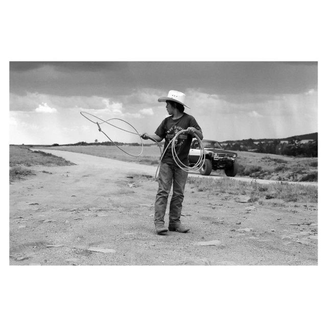Young person wearing a cowboy hat is holding a lasso rope on a dirt road with a vehicle and cloudy sky in the background.