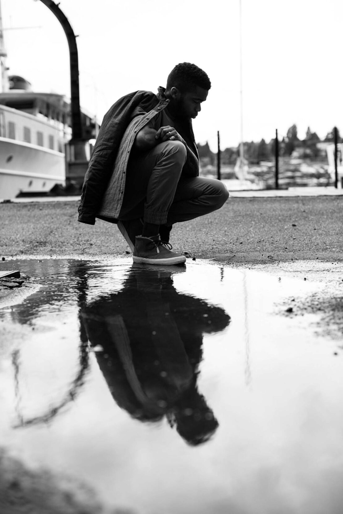 Monochrome image of an African American adult male crouching near a puddle reflecting his image; a boat and trees in the background.