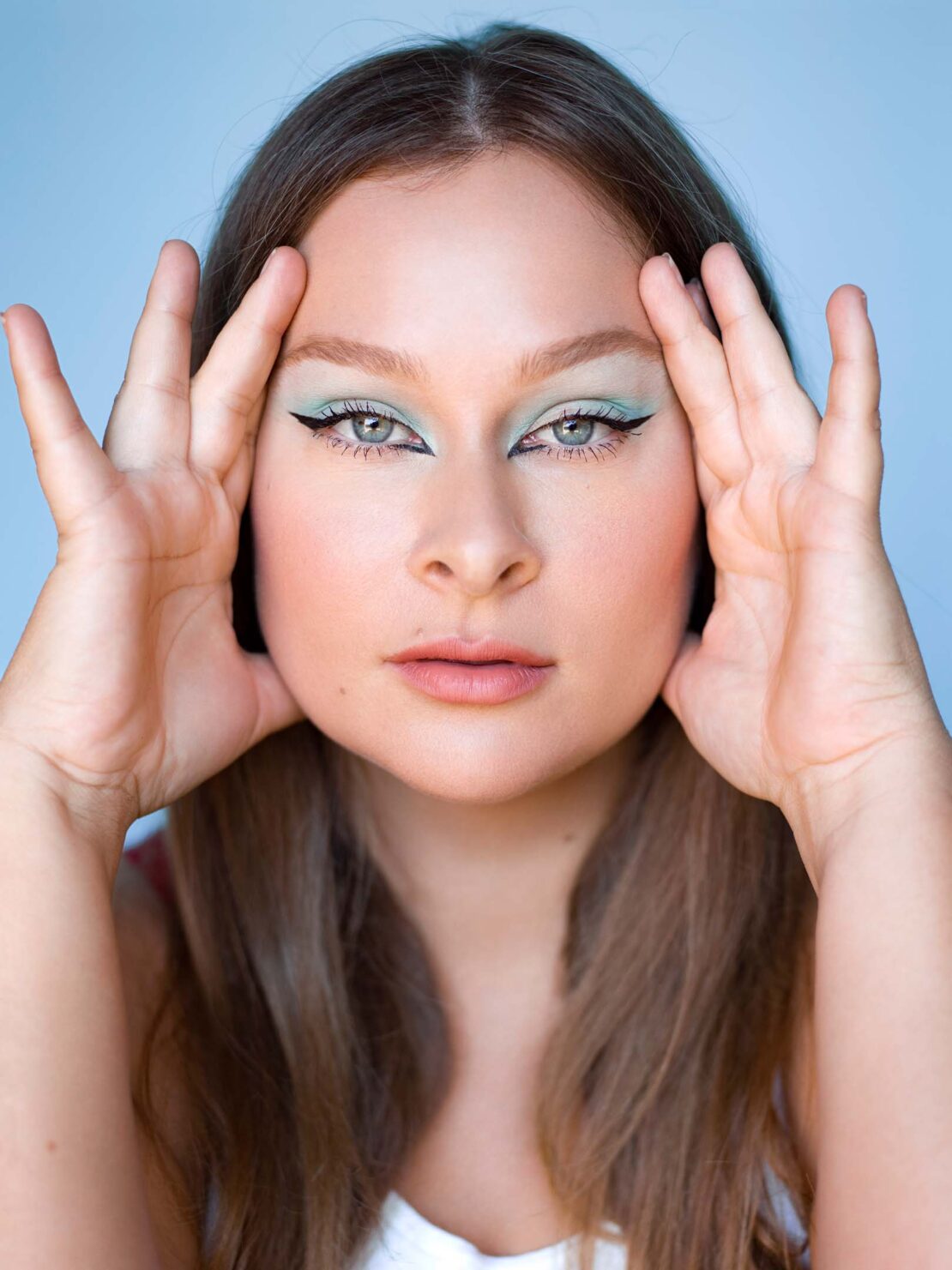 Close-up portrait of a young woman with creative makeup posing against a light blue background, showcasing her face and hairstyle.